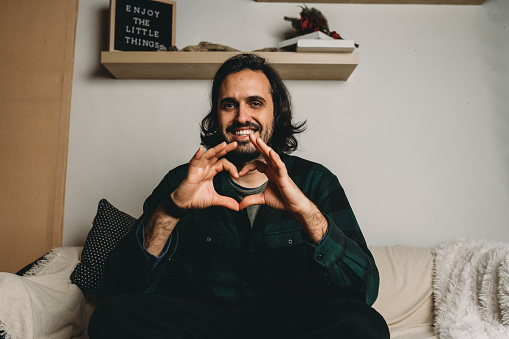 Portrait of a millennial man doing an heart shape to the camera. He's smiling, looking at camera.