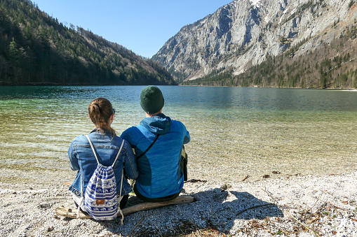 A couple sitting at the shore of Leopoldsteiner lake in Austria. The lake is surrounded by high Alps. The shallow water is crystal clear, spring water has a calm surface. Looking in the same direction