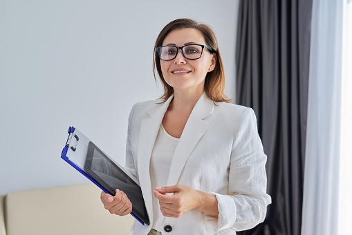 Business portrait in the office, confident smiling middle-aged businesswoman, female in glasses with tablet papers looking at camera