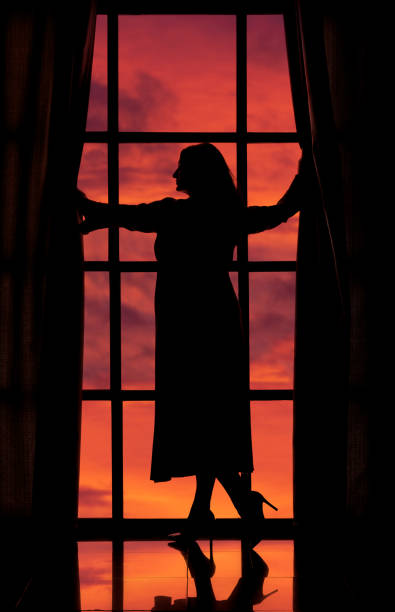 silhouette of a woman in a long peignoir by a large window against the background of an orange sunset sky beautifu silhouette of a woman in a long peignoir by a large window against the background of an orange sunset sky seduction stock pictures, royalty-free photos & images