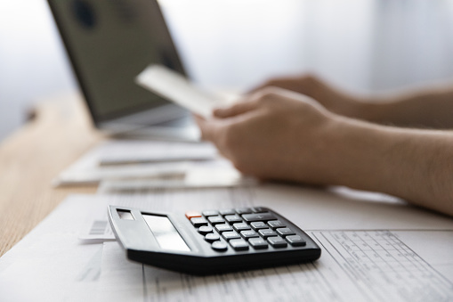 Crop close up of man sit at home desk work on laptop calculate household expenses expenditures on calculator. Male busy with paperwork document, manage house family finances or budget online.