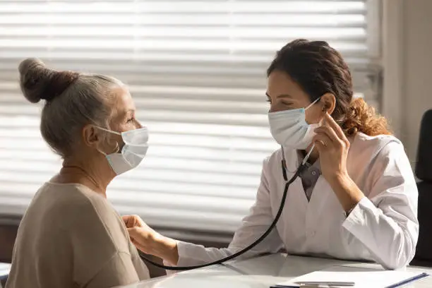 Caring young female doctor in medical facemask hold stethoscope listen to elderly patient heart in hospital. Woman GP or physician do regular checkup examine mature client at consultation in clinic.