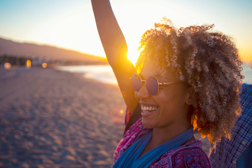 Woman dancing on the beach with friends at sunset or sunrise. She is having fun and smiling. She has an Afro and is of African decent. The beach and ocean can be seen in the background.