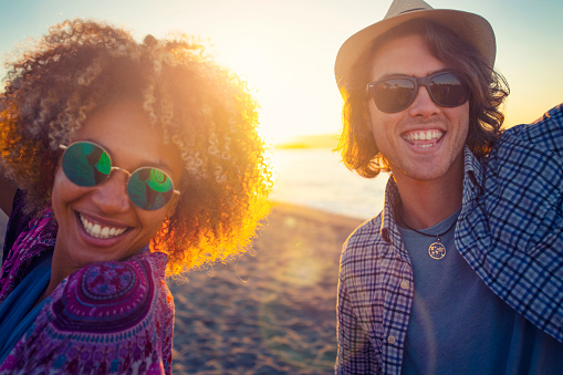 Couple dancing on the beach with friends at sunset or sunrise. They are having fun laughing and smiling. She has an Afro and is of African decent. He is Caucasian and wearing a hat. The beach and ocean can be seen in the background. looking at the camera