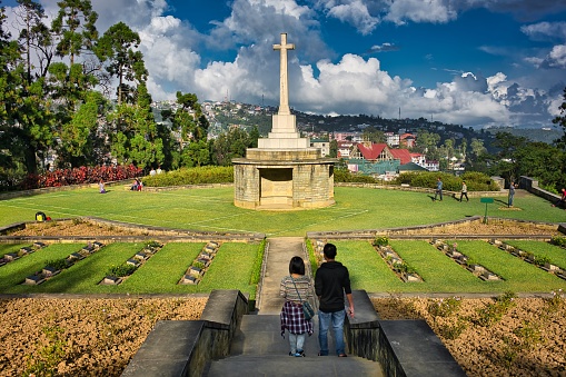 Kohima, India - October 30, 2016: Two people in front of the big white cross of Kohima War Cemetery, while other people take photos and admire the cemetery on a sunny day, in Nagaland region, in Seven Sisters of India.