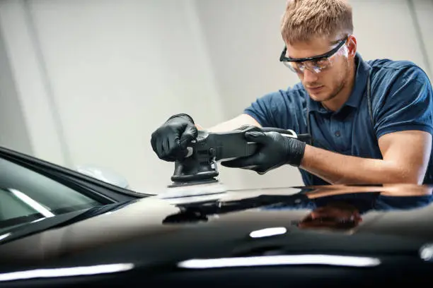 Closeup side view of a young man polishing driver's door of a black passenger car. He's using power polisher with wool brush.