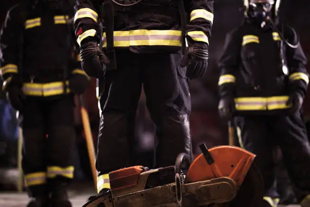 Group of abstract professional firefighters with full equipment, oxygen masks, and emergency rescue tools, circular hydraulic and gas saw, axe, and sledge hammer. Firetrucks in the background.