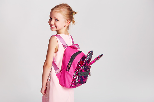 Cute little girl with bunny ears backpack in pink sequin dress. Childhood, education and people concept - happy kid girl with school bag over grey background
