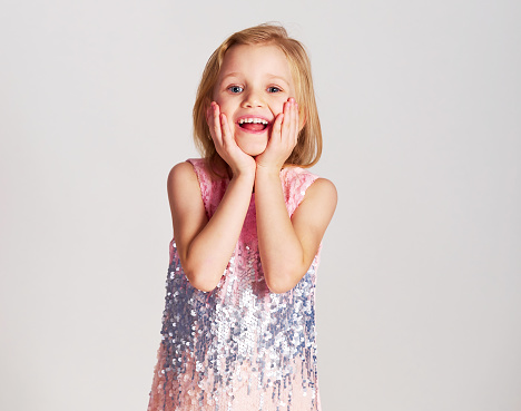 Surprised and happy young girl blond hair and blue eyes posing and fool around in fashionable pink sequin dress in studio on grey background. Sale, holidays, birthday party concept.