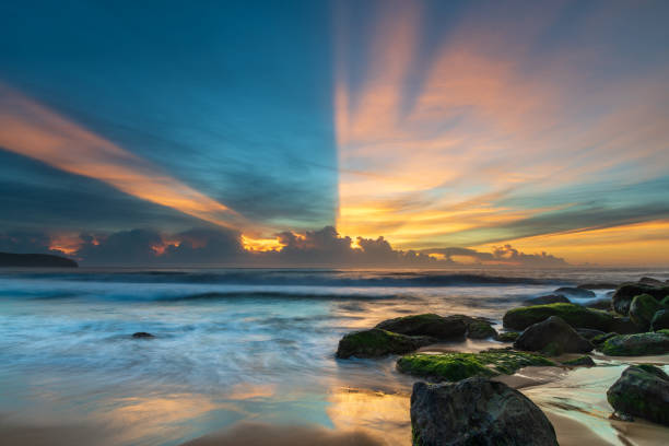 High clouds drift over a cloud bank on the horizon, sun rays streak over the sky creating a beautiful sunrise seascape from Killcare Beach on the Central Coast, NSW, Australia.
