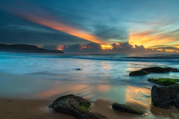 High clouds drift over a cloud bank on the horizon, sun rays streak over the sky creating a beautiful sunrise seascape from Killcare Beach on the Central Coast, NSW, Australia.