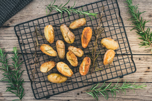 Baked unpeeled halfed potatoes with salt and rosemary on a black cooling rack on a rustic wooden table, top view