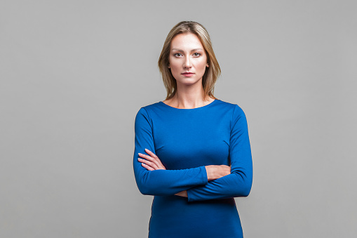 Portrait of young confident businesswoman in elegant tight blue dress standing with crossed hands, looking successful, smart and professional. indoor studio shot isolated on gray background