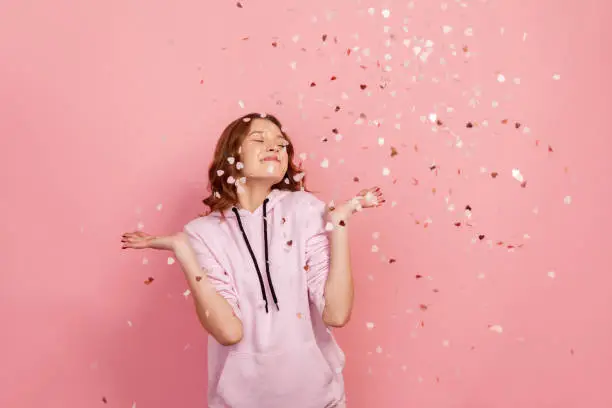 Photo of Extremely happy young brunette female enjoying falling heart shaped confetti and smiling with closed eyes, celebrating valentines day, holiday mood