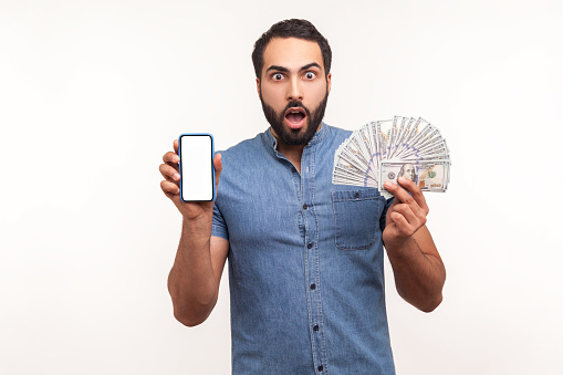 Surprised shocked man holding empty screen smartphone ad fan of hundred dollars, looking at camera with astonishment, banking and earning online. Indoor studio shot isolated on white background