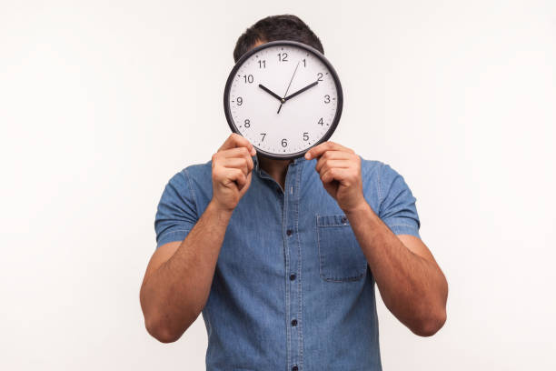 istock Man in blue shirt hiding face behind wall clock display, wasting his time, procrastination, organization of working time 1294803755