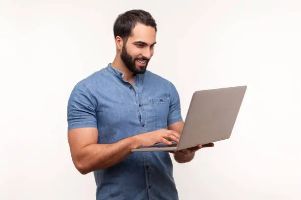 Smiling positive man holding laptop in hand and typing, blogger making posts in social networks, chatting with followers. Indoor studio shot isolated on white background