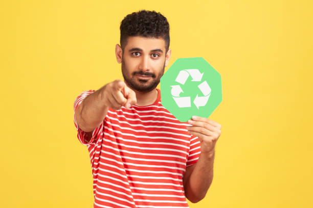 hombre barbudo serio sosteniendo el cartel de reciclaje verde y apuntando con el dedo mirando a la cámara, pidiendo la clasificación de basura y la protección del medio ambiente - recycling recycling symbol environmentalist people fotografías e imágenes de stock