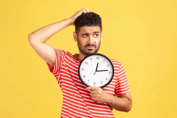 Photo of Confused thoughtful man with beard in red striped t-shirt rubbing his head holding in hands big wallclock, time out, worried about deadline