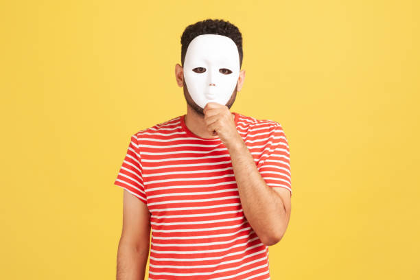 Unknown anonymous man in striped red t-shirt covering his face with white mask, hiding personality, conspiracy and privacy, secrets. Unknown anonymous man in striped red t-shirt covering his face with white mask, hiding personality, conspiracy and privacy, secrets. Indoor studio shot isolated on yellow background hypocrisy stock pictures, royalty-free photos & images
