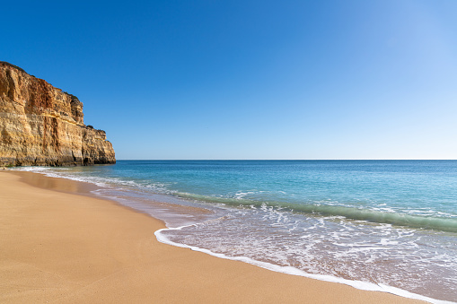 An empty golden sand beach wth tropical turquoise water and rocky cliffs under a blue sky