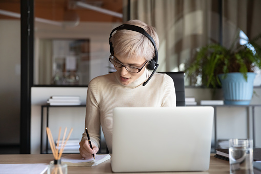 Businesswoman wearing headset writing notes, using laptop in office, female employee intern watching webinar, studying, manager consulting client online, making video call, internet negotiations