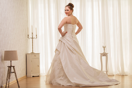 Rear view of beautiful young bride in a puffy white wedding dress standing with her hands on hips and smiling at camera with confidence.