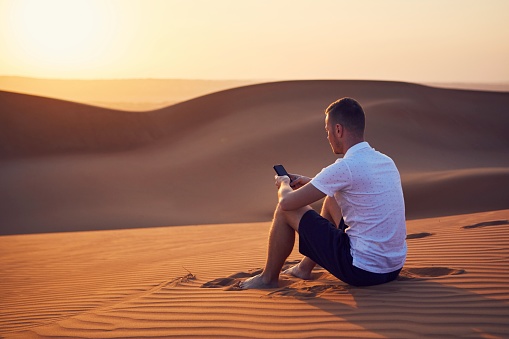 Alone in desert. Young man sitting on sand dune and using phone at golden sunset. Wahiba Sands in Oman.
