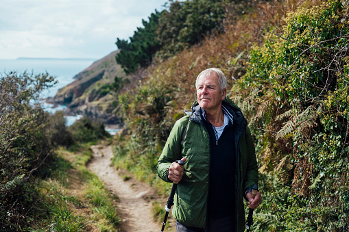 A senior man using hiking poles, on a hike on a coastal path along the coastline of Cornwall. He is looking away from the camera at the view.