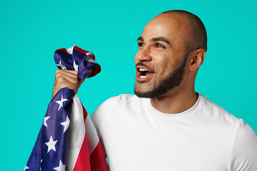 One person of aged 30-39 years old caucasian young male standing in front of white background wearing shirt who is shouting and celebrating and showing patriotism and holding flag