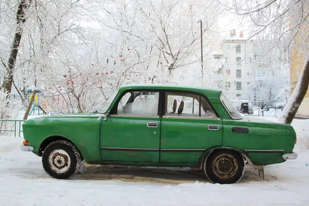 A old green russian car, a moskvich, is standing in the yard of house in wintertime; a lot of snow an rowan berries surround it; Kaluga, Russia