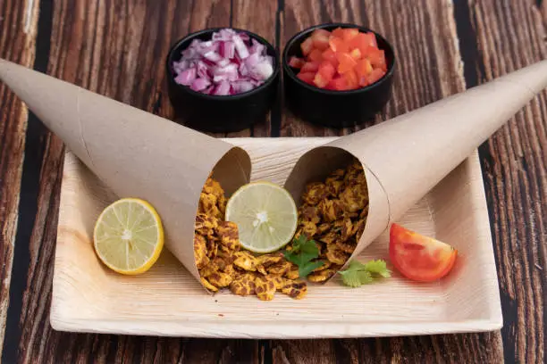 Photo of Chatpata Namkeen Masala Chana Jor Zor Garam In Traditional Brown Colored Paper Cone Served With Lemon And Tomatoes And Sliced Onions In Eco Friendly Tray. Brown Wooden Textured Background