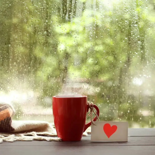 red mug with a note depicting a heart, on the table by the window with drops after rain