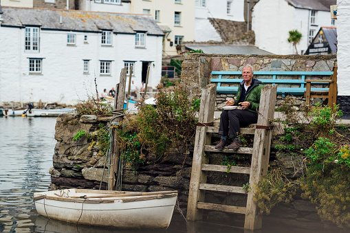A senior man sitting on a wooden ladder at the harbour of Polperro, Cornwall. He is eating a polystyrene tray of chips while overlooking the water.