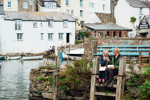 A senior man and his wife sitting on a wooden ladder at the harbour of Polperro, Cornwall. They are eating a polystyrene tray of chips and laughing with each other.