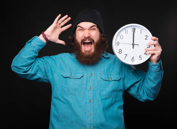 istock A stressed young bearded man is holding a big white clock  and is looking very stressed at the camera near a black wall 1294893266