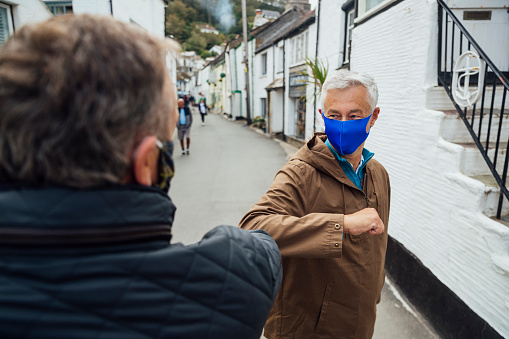 Over the shoulder view of two men wearing protective face masks greeting each other with an elbow bump. They are doing this to try reduce the spread of germs during the COVID-19 pandemic.