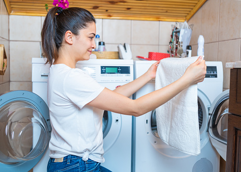 Young woman is collecting towels and clothes from laundry tumble dryer in bathroom.  Laundry day