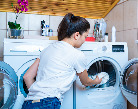 Young woman is collecting towels and clothes from laundry tumble dryer in bathroom.  Laundry day