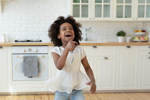 Overjoyed little African American girl holding fork as microphone and singing, standing in modern kitchen, having fun with kitchenware at home, cute child playing funny game, listening to music
