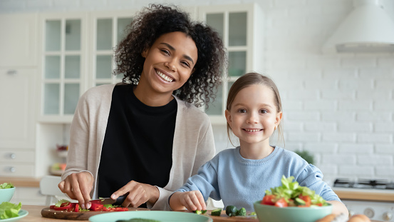 Head shot portrait smiling African American woman with adopted Caucasian daughter cooking salad together, sitting at wooden table in kitchen, multiracial family preparing meal, enjoying weekend