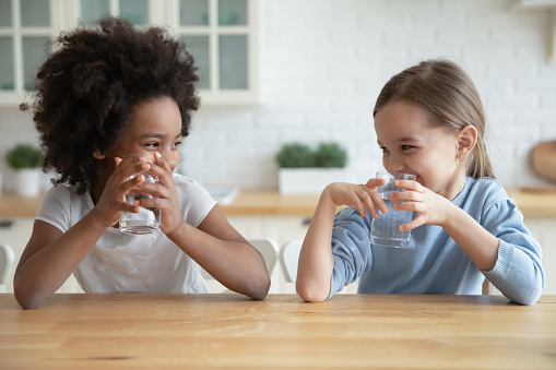 Cute smiling diverse little girls drinking fresh water, sitting at wooden table in kitchen, looking at each other, multiracial sisters adorable kids enjoying aqua, refreshment concept