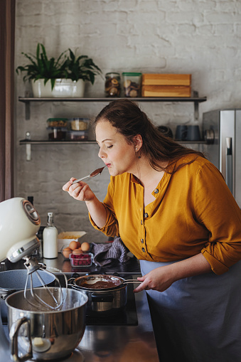 Beautiful young happy Caucasian woman tasting a hot chocolate sauce over a kitchen stove while making a cake at home.