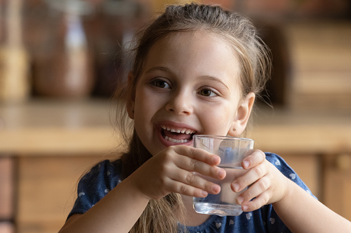Enjoying fresh water. Close up of happy little girl feeling glad drinking cool clear natural mineral aqua without gas from glass. Small kid looking aside with smile feeling healthy quenching thirst