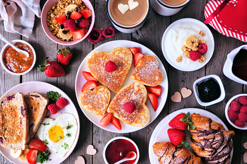 Valentines or Mothers Day brunch table scene. Top view on a dark wood background. Heart shaped pancakes, eggs and an assortment of love themed food.