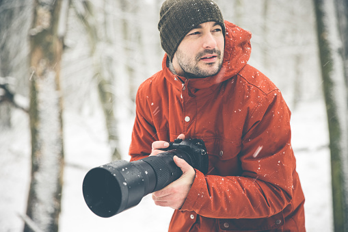Male wildlife photographer with long zoom lens on digital camera is on a photoshoot in a snow blizzard during winter in Denmark. The photographer wears a green knit hat and a red winter jacket that pops in the white winter wonderland.