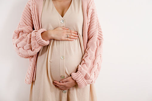 Young beautiful woman on second trimester of pregnancy. Cropped shot of pregnant female in pink oversized knitted sweater with arms on belly. Expecting a child concept. Isolated background, copy space