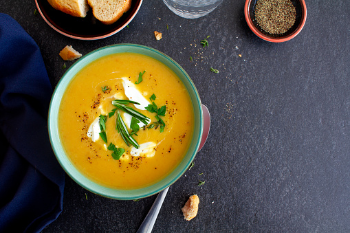 A hearty pumpkin and vegetable soup, with cream sprinkled with fresh herbs and black pepper. Served with fresh crusty bread, shot from above on a grey slate background. Flat lay. \nCopy space to the right of the image.