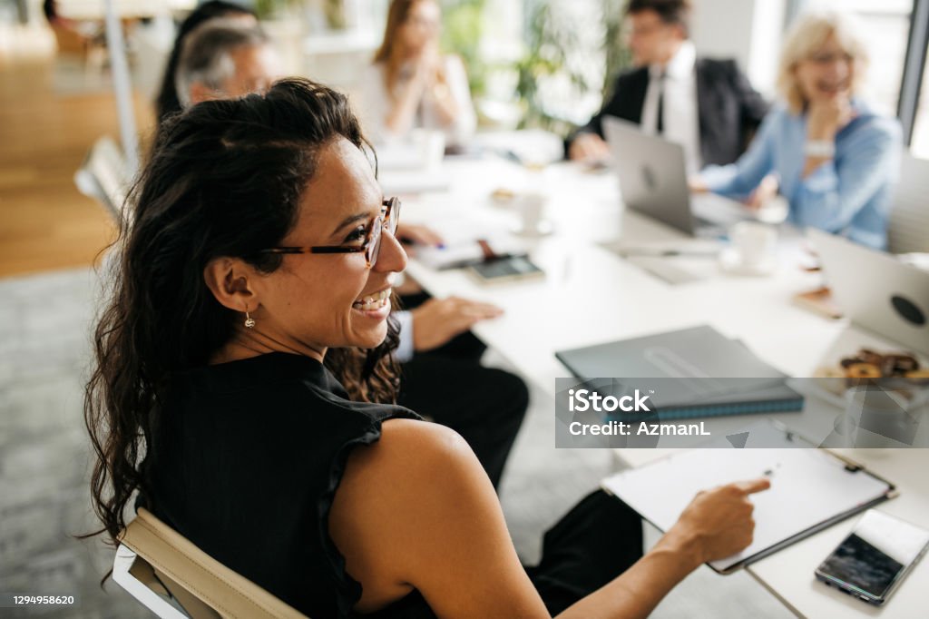 Candid Close-Up of Hispanic Businesswoman in Office Meeting Over the shoulder profile of bespectacled female executive in early 30s sitting at conference table and laughing as she interacts with off-camera colleague. Office Stock Photo