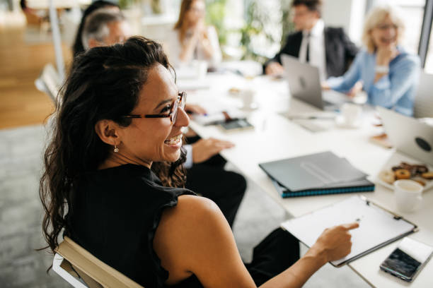 Candid Close-Up of Hispanic Businesswoman in Office Meeting Over the shoulder profile of bespectacled female executive in early 30s sitting at conference table and laughing as she interacts with off-camera colleague. conference event photos stock pictures, royalty-free photos & images
